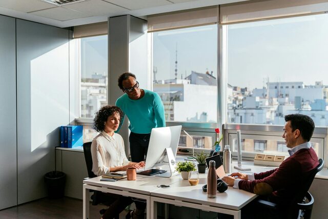 Three colleagues discussing work at a desk in a sunlit office